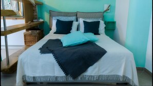 Een net opgemaakt bed met een wit en donkerblauwe dekbedovertrek, twee kussens en een blauwe plaid. Er is een houten plank met manden en een rieten mand op de vloer naast het bed.