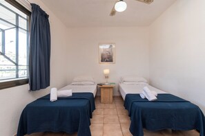 Ein Zimmer mit zwei Betten, einem Nachttisch aus Holz mit Lampe, einem gerahmten Bild an der Wand und einem Fenster mit Vorhängen.