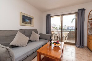 Ein Wohnzimmer mit grauem Sofa, einem Holztisch mit Fruchtschale und Weinflasche, sowie einem Balkon mit Blick auf Gebäude und Palmen.