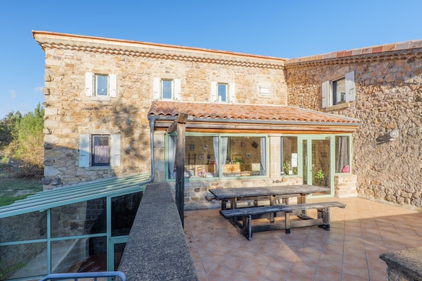 A stone building with a tiled roof, a glass-enclosed patio area, and a wooden table.