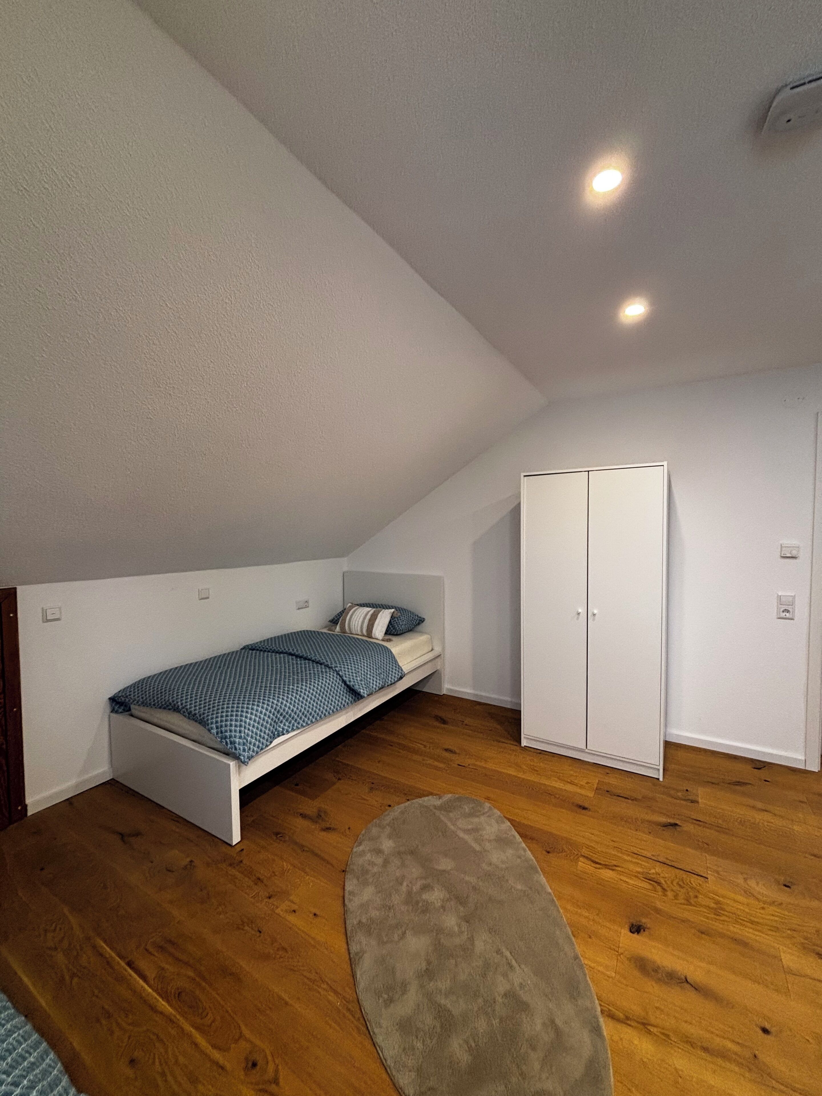 Una piccola camera con un letto singolo, soffitto spiovente, pavimento in legno, un armadio e un tappeto.
