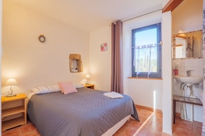 A bedroom with a bed, bedside tables, a window with curtains, a bathroom with a sink and mirror, and a wall-mounted decoration. - Guest Room “Rosée” with Shared Terrace, Communal Garden and Wi-Fi (Laurac-en-Vivarais)