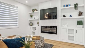A modern living room with a flat-screen TV mounted above a fireplace, white cabinetry, and decorative shelves. - Indoor Fireplace • 3 Kings • Free Parking • Desk (Calgary)