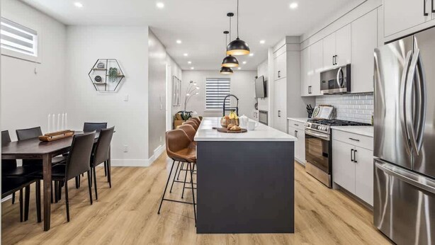 A modern kitchen with a central island, stainless steel appliances, and a dining area with a wooden table and chairs. - Indoor Fireplace • 3 Kings • Free Parking • Desk (Calgary)