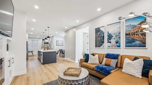 A modern living room with a brown leather sofa, a round coffee table, and a kitchen in the background. - Indoor Fireplace • 3 Kings • Free Parking • Desk (Calgary)