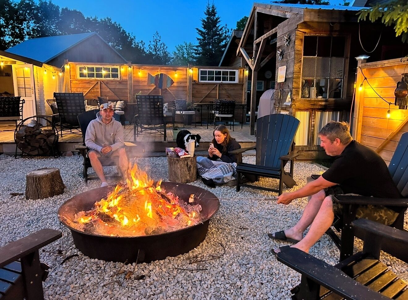 A cozy outdoor seating area with a fire pit, wooden chairs, and a rustic building in the background.