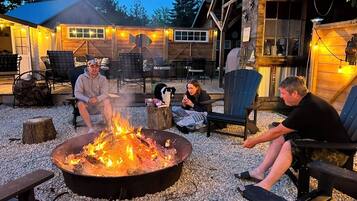 A cozy outdoor seating area with a fire pit, wooden chairs, and a rustic building in the background.