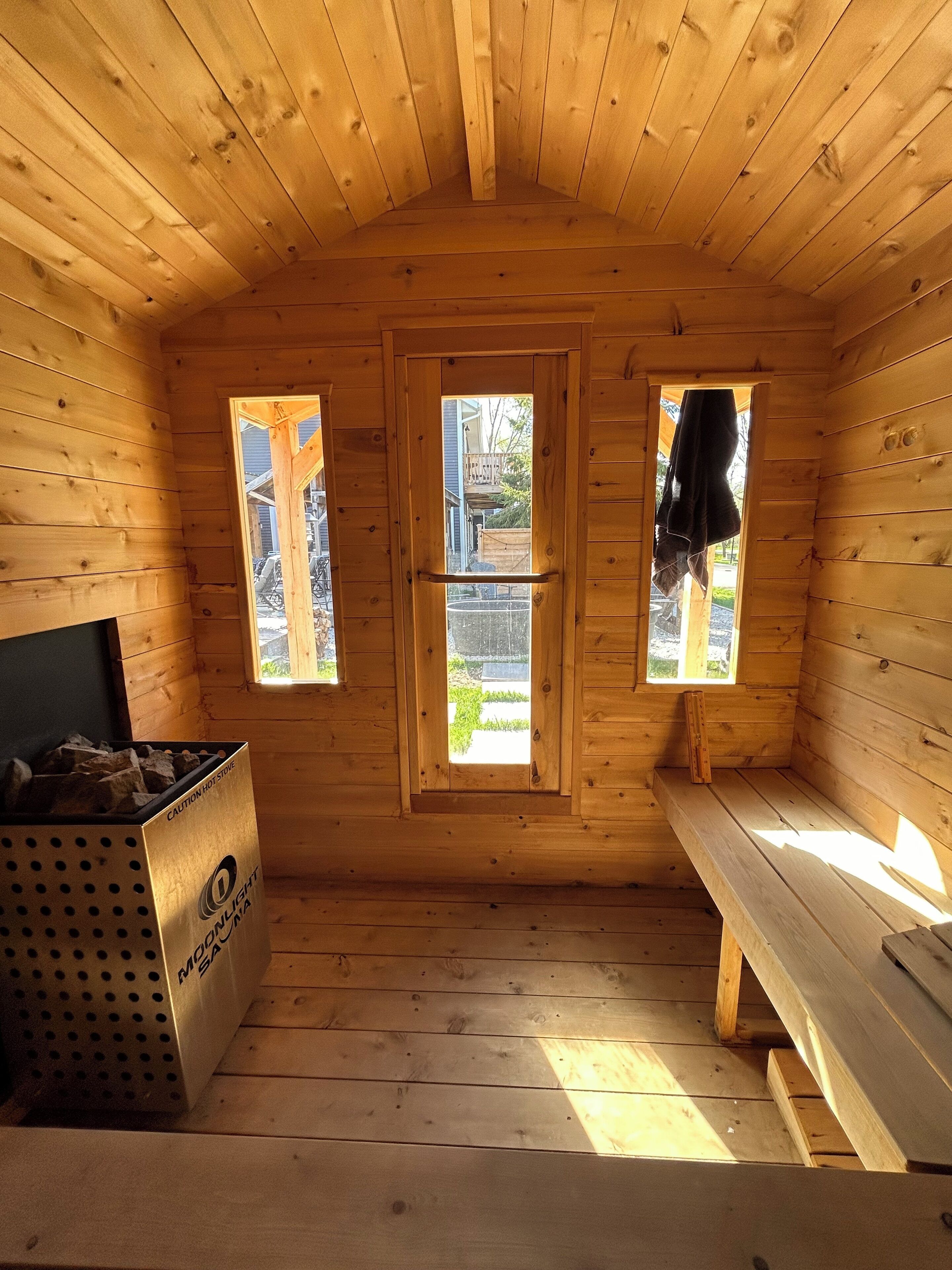 Une cabane en bois avec deux fenêtres, un banc et un récipient en métal portant l’inscription « ATTENTION, POÊLE CHAUD ».