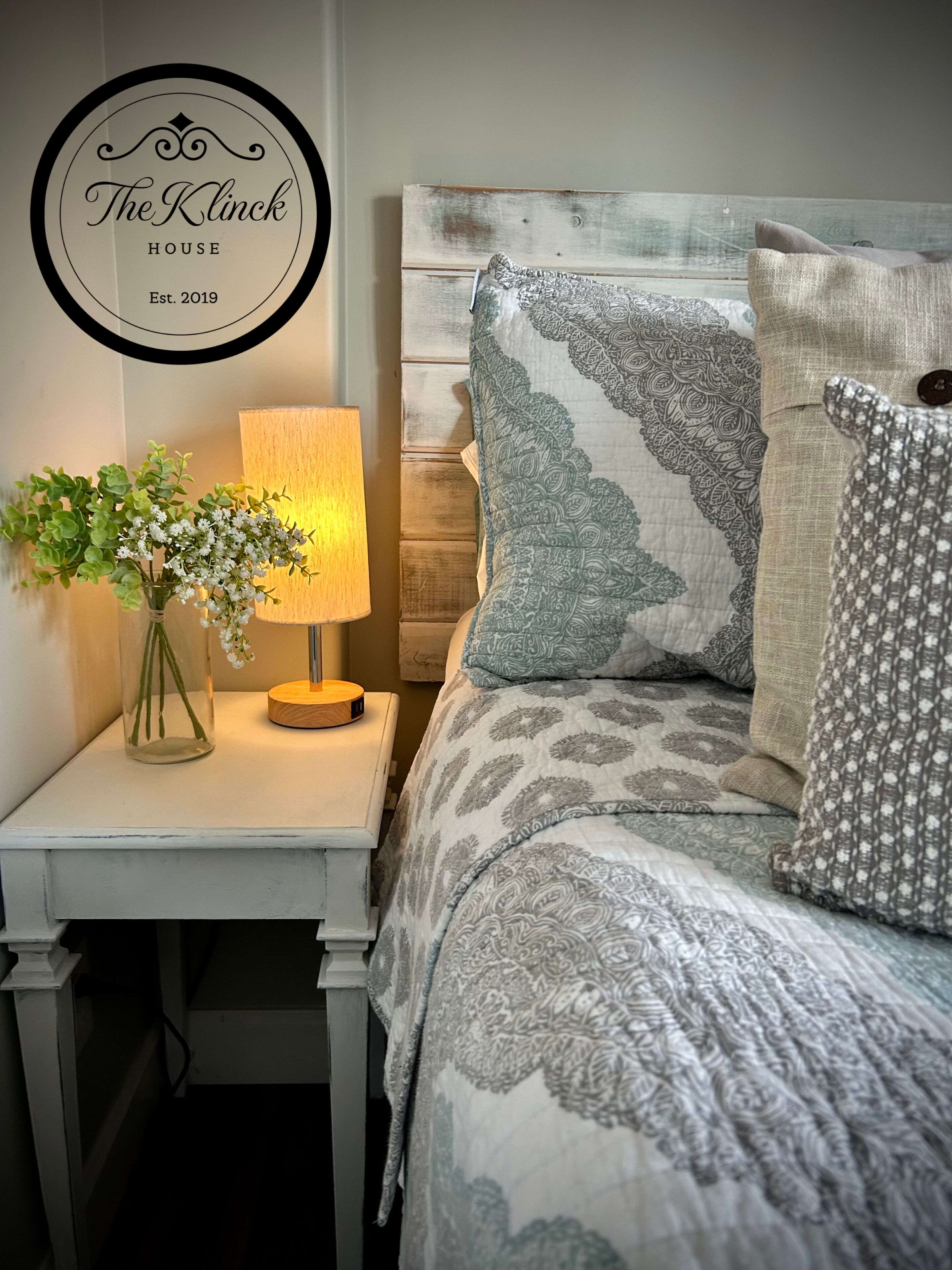 Un dormitorio ordenado con cama, almohadas, una mesita de noche con lámpara y un cartel en la pared.