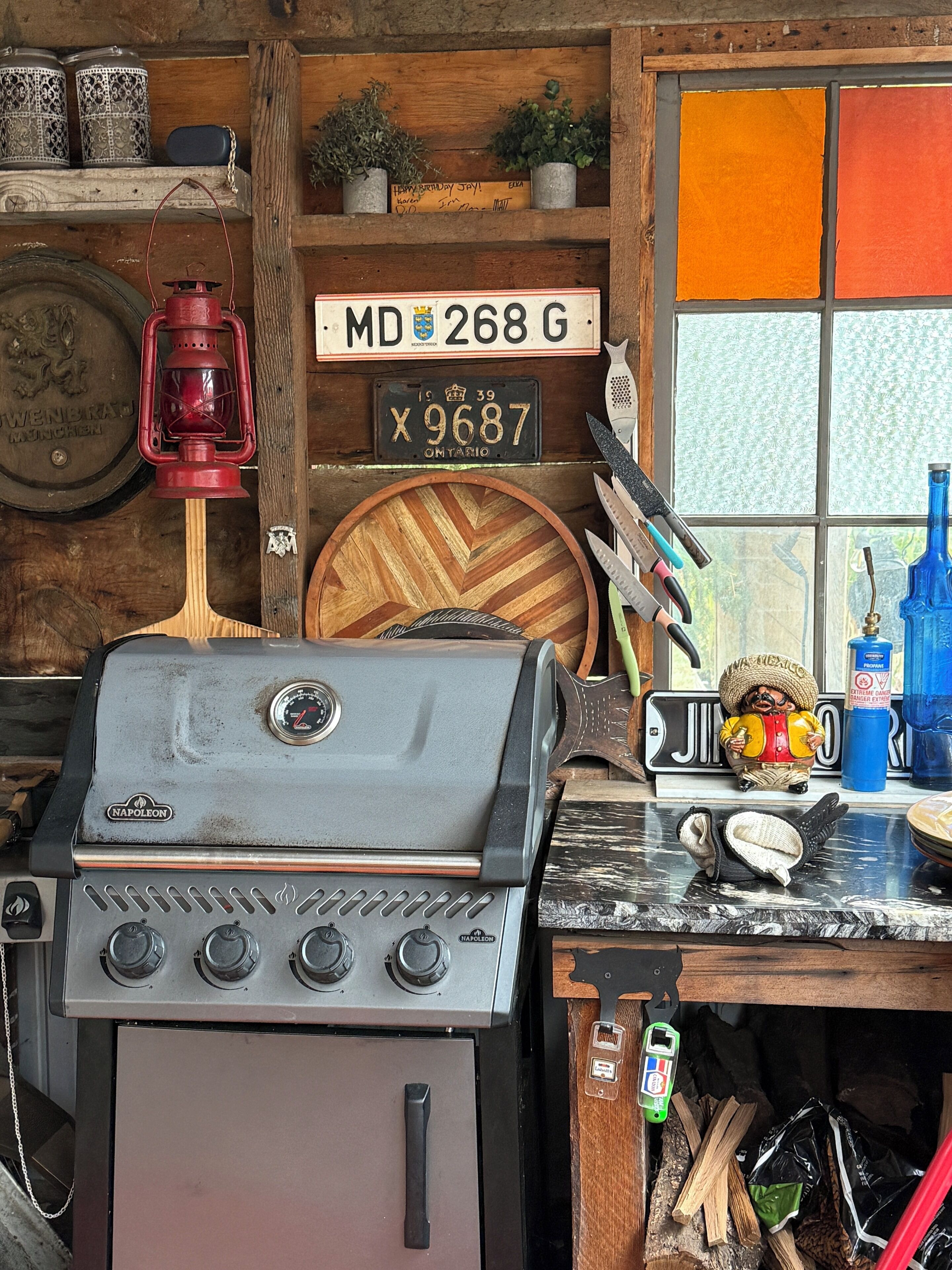 A kitchen with a Napoleon grill, wooden shelves with decorative items, a license plate, and a window with a view.