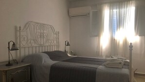 A bedroom with a bed, bedside tables, lamps, a window with curtains, and an air conditioning unit. - Apartment 'Cozy  For Rent In Kythera' with Shared Terrace, Wi-Fi and Air Conditioning (Κύθηρα)