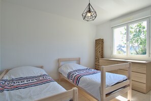 A bedroom with two wooden beds, a wooden dresser, a geometric pendant light, and a window with a view of greenery. - Holiday Home "Gîte with Game Room" with Private Terrace, Garden & Wi-Fi (Montfarville)