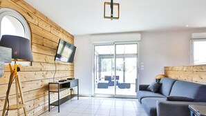 A modern living room with a wooden wall, a flat-screen TV, a sofa, a lamp, and a sliding glass door leading to an outdoor seating area. - Holiday Home "Gîte with Game Room" with Private Terrace, Garden & Wi-Fi (Montfarville)