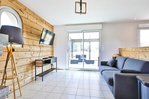 A modern living room with a wooden wall, a flat-screen TV, a sofa, a lamp, and a sliding glass door leading to an outdoor seating area. - Holiday Home "Gîte with Game Room" with Private Terrace, Garden & Wi-Fi (Montfarville)