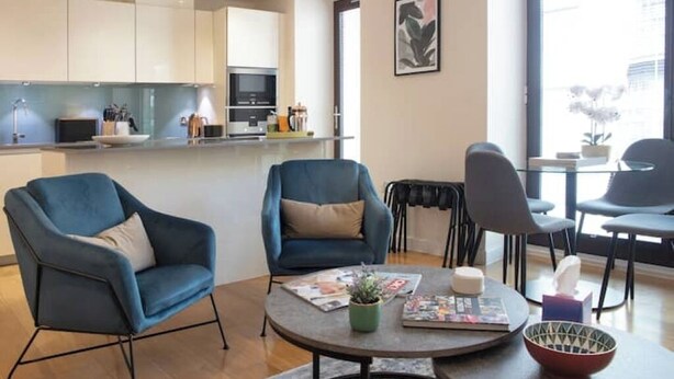 Ein modernes Wohnzimmer mit zwei blauen Sesseln, einem Couchtisch mit Zeitschriften und einer Pflanze, sowie einer Küche im Hintergrund.