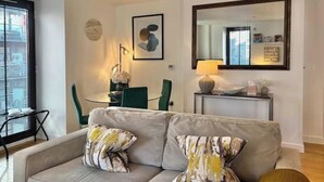 Una sala de estar con un sofá, dos almohadas, una mesita de noche con una lámpara, un cuadro enmarcado y un espejo en la pared.