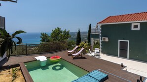 Una piscina en la azotea con sillones de descanso, vista al mar y un edificio verde con techo rojo.
