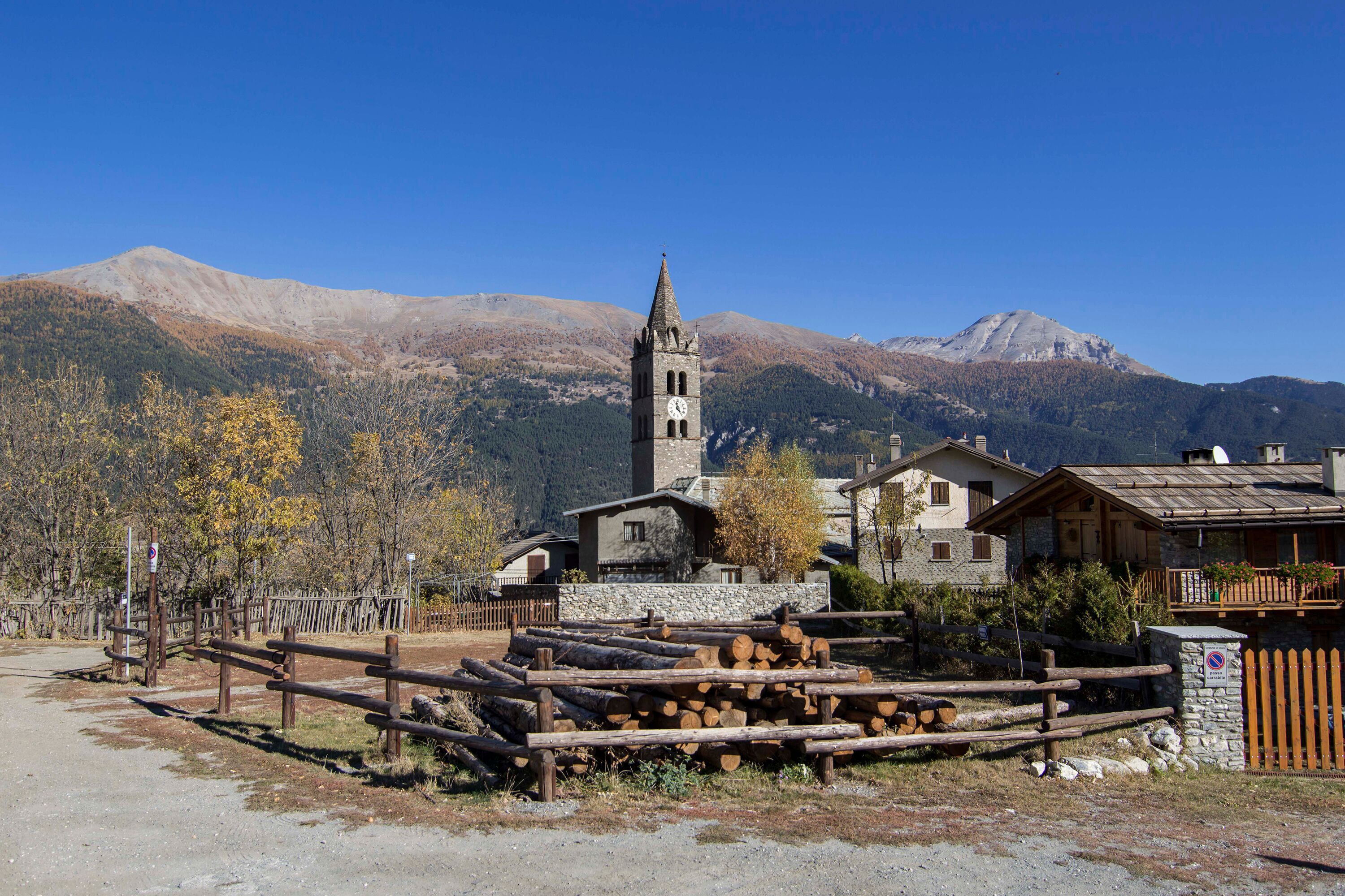 A rustic mountain village with a church featuring a clock tower, surrounded by wooden houses and a clear view of mountains in the background.