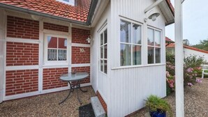 Il s’agit d’un petit bâtiment d’un seul étage, doté d’un toit recouvert de tuiles rouges, d’une façade en briques et d’un mur extérieur peint en blanc. On y trouve une petite table et une plante en pot près de l’entrée.