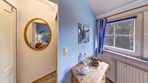 Une pièce avec une commode en bois, un miroir rond, une fenêtre avec des rideaux bleus et un radiateur.