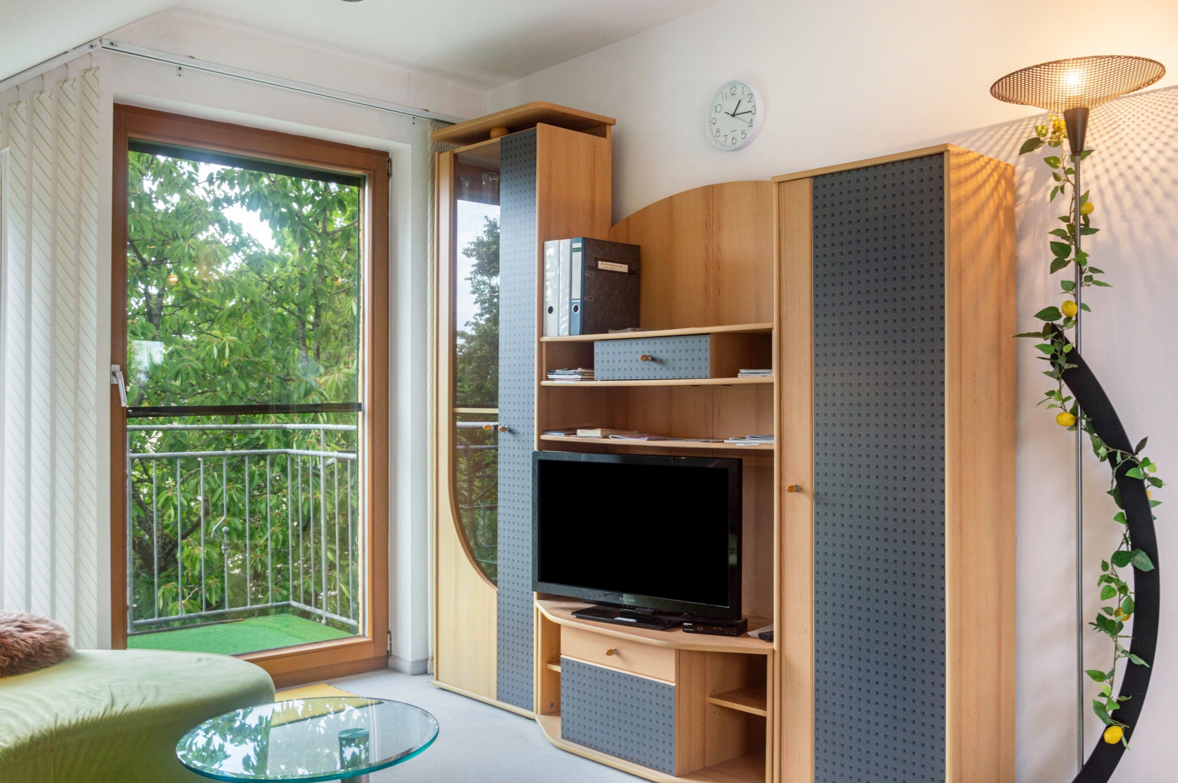 A modern living room with a TV, storage cabinets, a glass coffee table, and a balcony with a view of greenery.