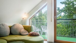 Una habitación con sofá verde, sillas blancas, un ventanal con vista a zonas verdes y una lámpara de pie.