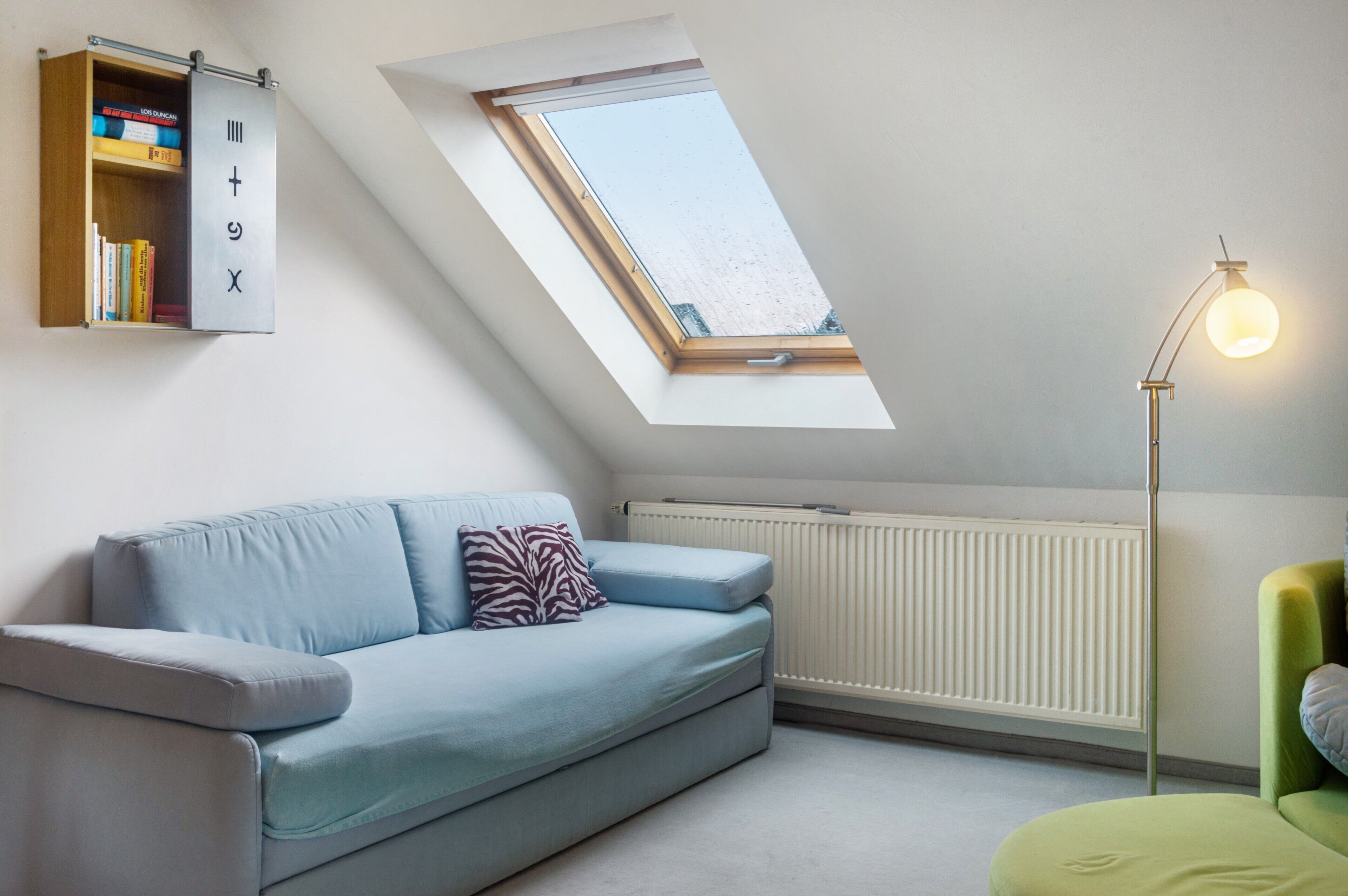 A room with a light blue sofa, a green armchair, a skylight, a radiator, and a floor lamp.