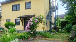 Un edificio amarillo con una escalera, un jardín con diversas plantas y un sendero pavimentado.