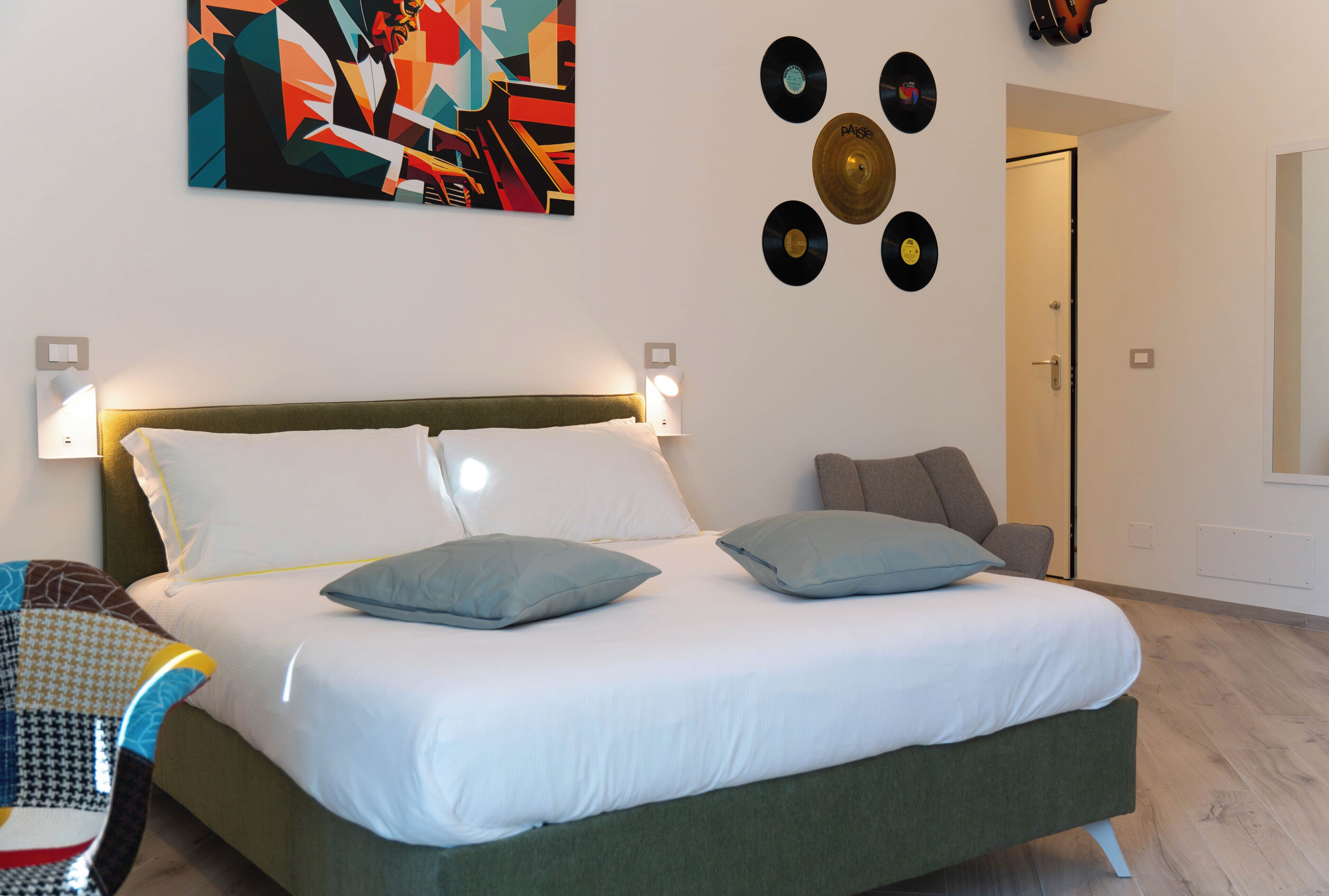 A modern bedroom with a green bed, white linens, and a colorful abstract painting above the bed. There are vinyl records on the wall and a chair with a patterned cushion.