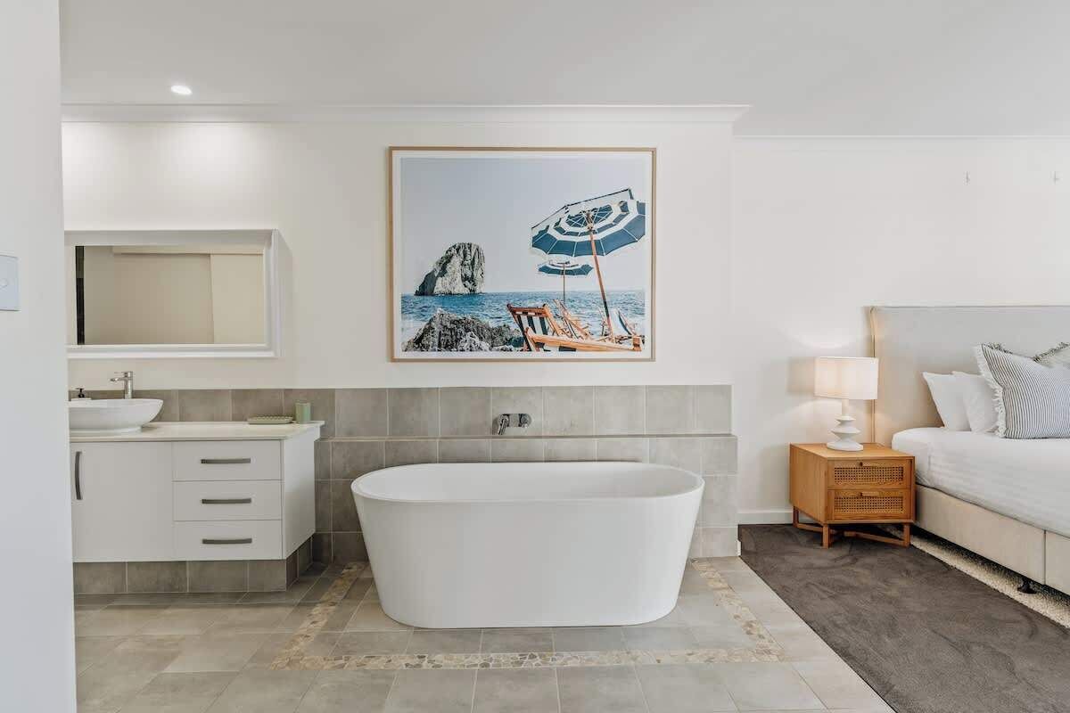 A modern bathroom with a freestanding bathtub, a large mirror, a framed picture of a beach scene, and a bedside table with a lamp.