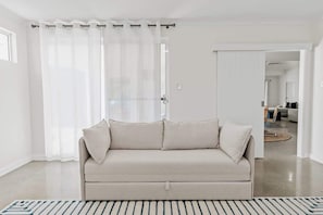 A neutral-toned sofa in a room with a striped rug, a curtained window, and a visible doorway leading to another room. - Sundrift—Sophisticated Beachside Retreat with Pool (Quindalup)