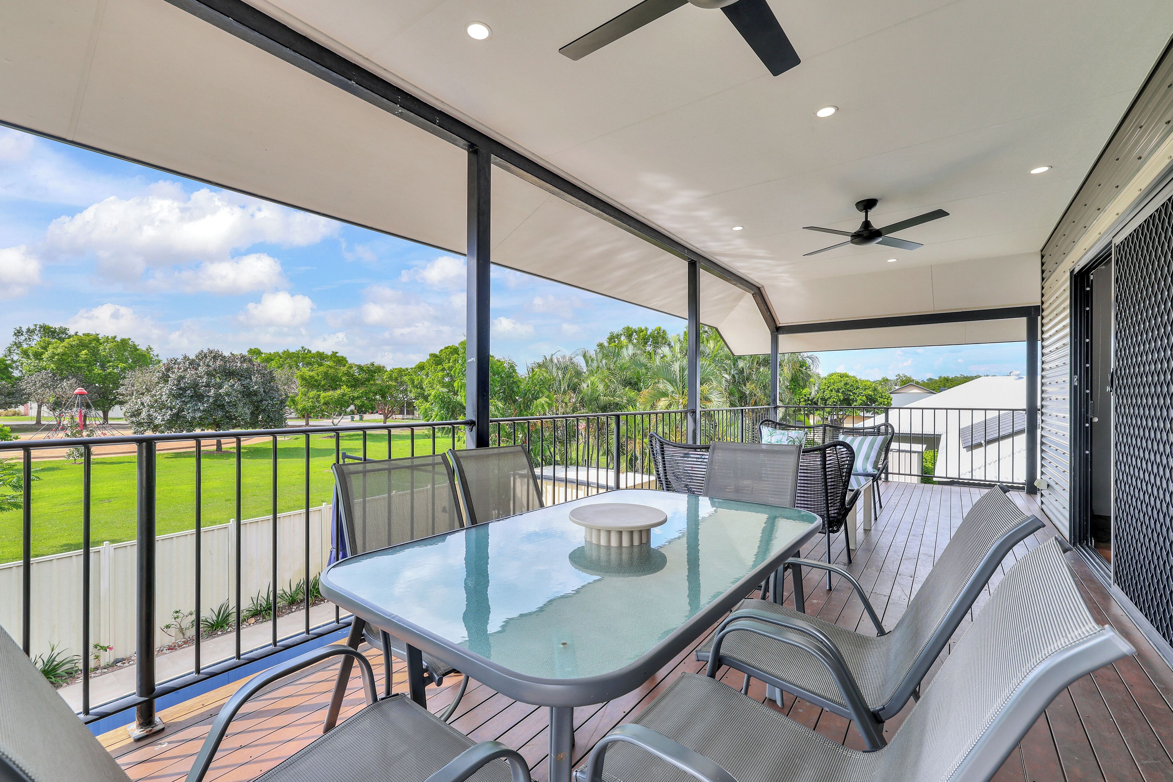 A covered patio with a glass-top table and metal chairs, overlooking a green lawn and trees.
