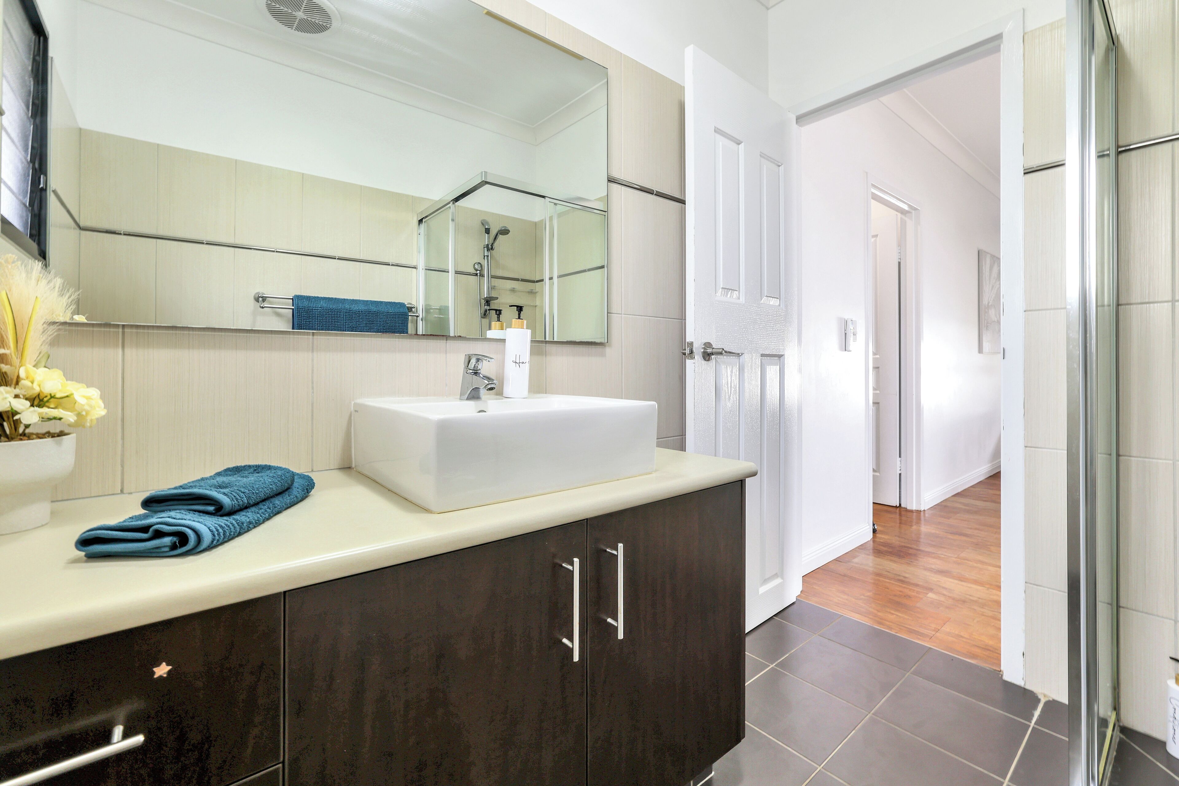 Un baño moderno con un espejo grande, lavamanos blanco, armarios oscuros y una cabina de ducha de vidrio.