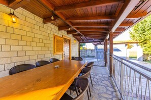 A covered outdoor dining area with a long wooden table and chairs, brick walls, and a stone floor.