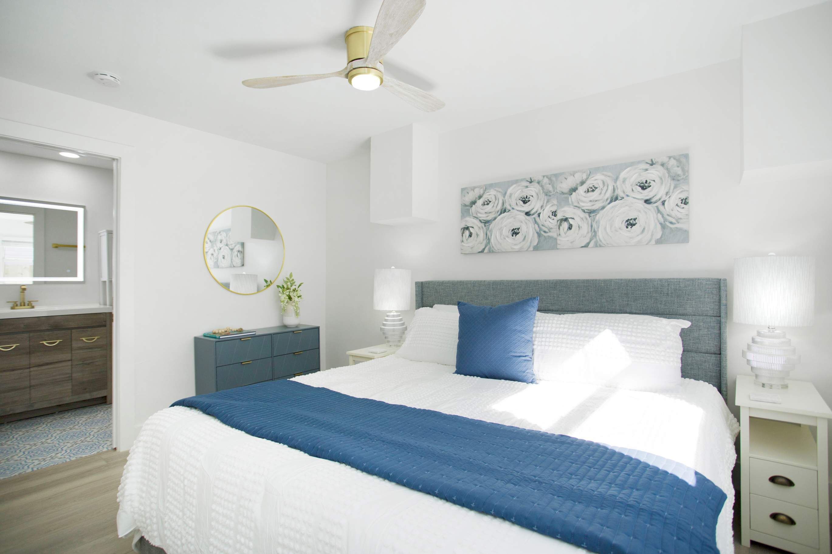 A modern bedroom with a large bed, a ceiling fan, a bedside table with a lamp, a dresser, and a wall-mounted mirror.