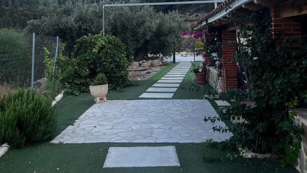 Un chemin mène à un bâtiment dont le toit est recouvert de tuiles, entouré de verdure et avec une montagne en arrière-plan.