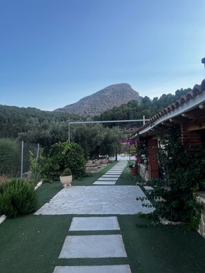 A pathway leading to a building with a tiled roof, surrounded by greenery and a mountain in the background.