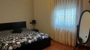 Une chambre à coucher avec un lit, une table de chevet, une lampe, un miroir et une fenêtre avec des rideaux.