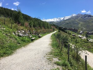 Uma trilha de cascalho serpenteia por uma região montanhosa, com vista para picos nevados ao longe.