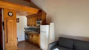 Een keuken met houten kastjes, een magnetron en een koelkast. In de woonruimte staat een rieten stoel en een grijze bank.