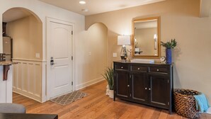 A hallway with wooden flooring, a white door, a dark wooden cabinet with a mirror, a lamp, and a potted plant. - Beautiful Plaza Condo | 01 (Kansas City)