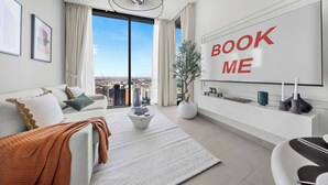 A modern living room with a large window, a sofa, a coffee table, a TV wall with 'BOOK ME' text, and a city view. - StayShort - Contemporary 1BR Meydan Sleeps 4 (Meydan)