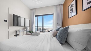 A modern hotel room with a large bed, a flat-screen TV mounted on the wall, a small side table with a plant, and a view of the city through large windows. - StayShort - Contemporary 1BR Meydan Sleeps 4 (Meydan)