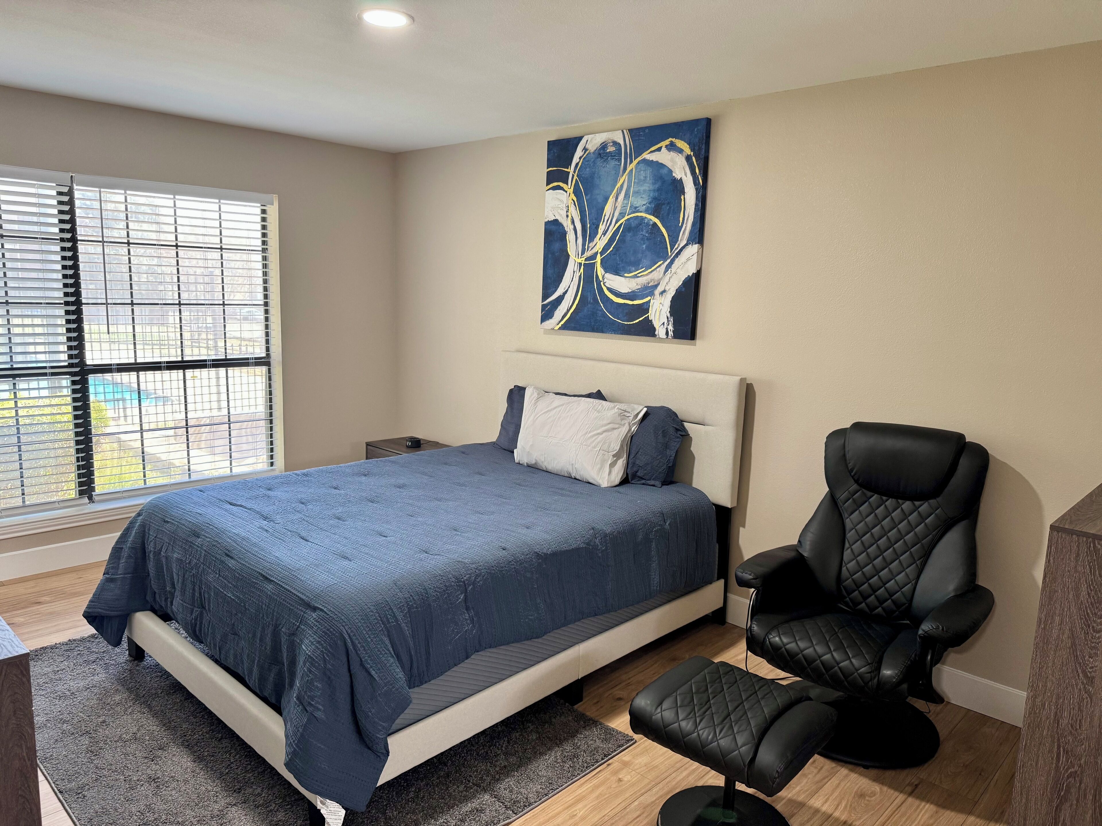 Een slaapkamer met een bed, een zwarte leren fauteuil, een nachtkastje en een schilderij aan de muur.