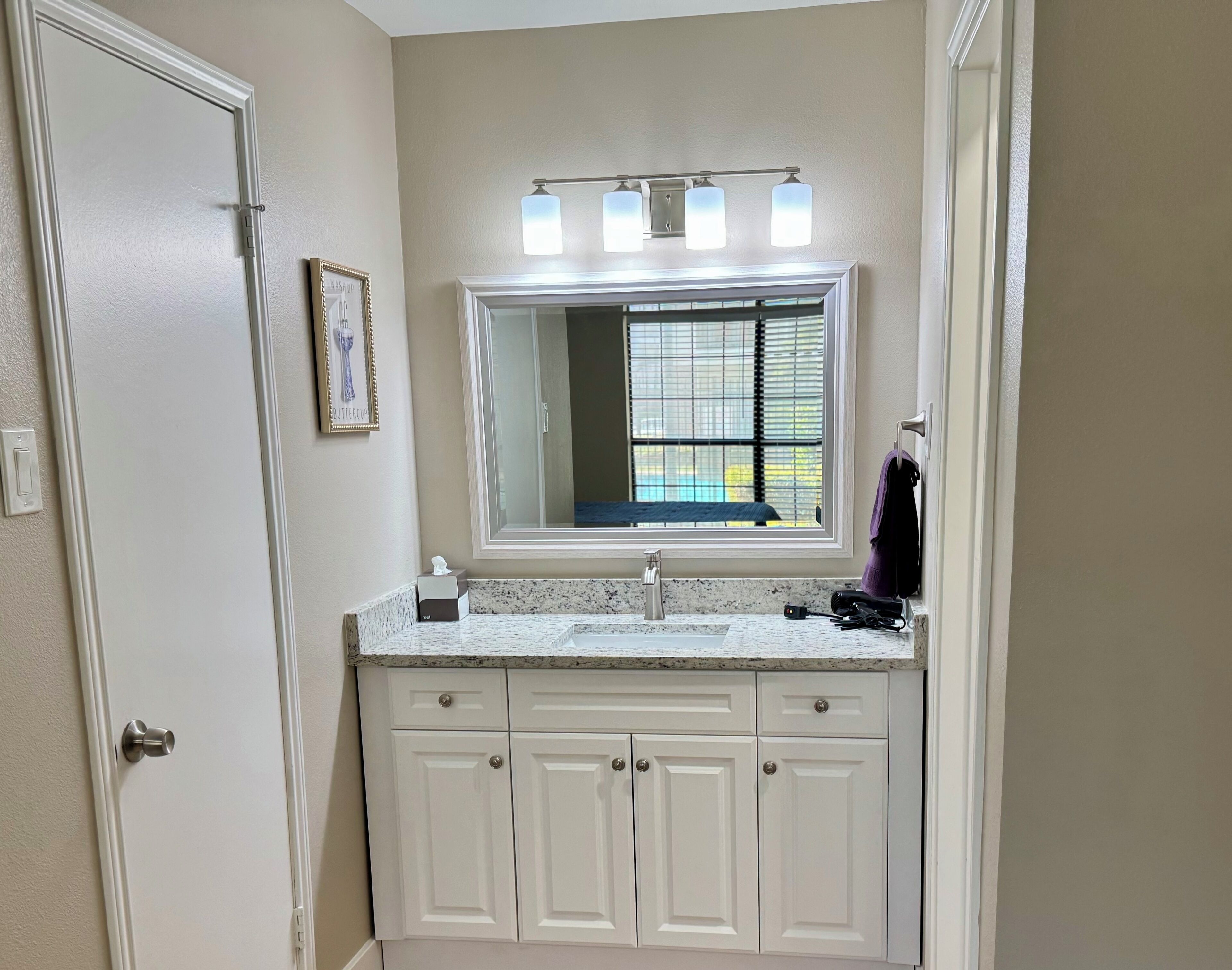 A bathroom with a large mirror, a vanity with a countertop, and a window with blinds.