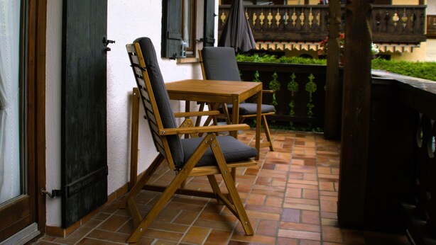 Une terrasse avec une table en bois et deux chaises, un luminaire fixé au mur et une vue sur un immeuble doté d’un balcon.