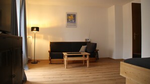 Une pièce avec un plancher en bois, un canapé noir, une table basse en bois, un lampadaire et une affiche accrochée au mur.