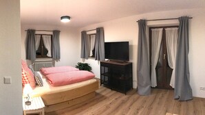Une chambre à coucher avec un lit, une télévision, un banc en bois et une fenêtre avec des rideaux.