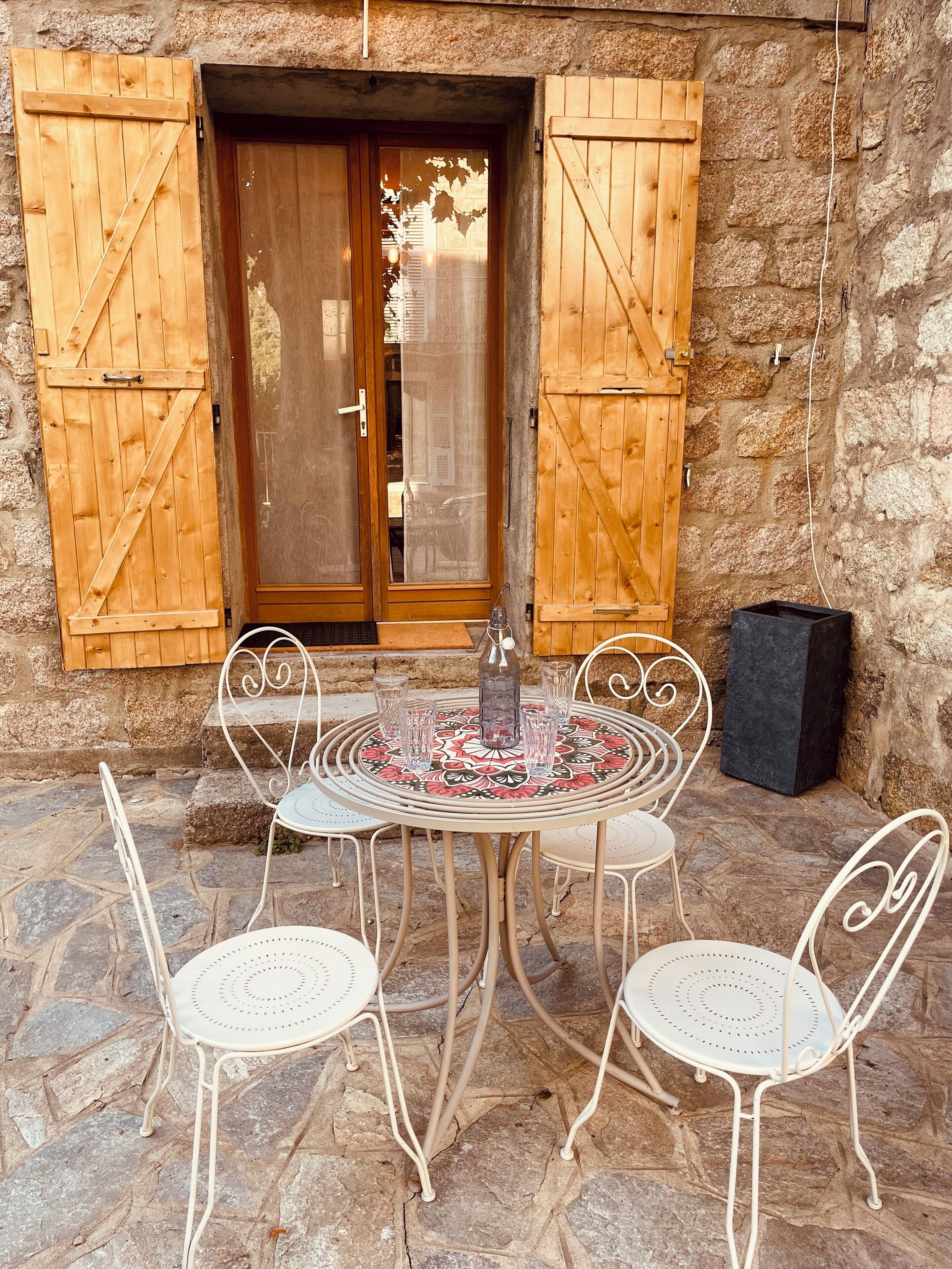 An outdoor seating area with a round table and four chairs, a bottle, and glasses on a patterned tablecloth.