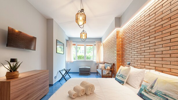 A modern hotel room with a large bed, a flat-screen TV, a wooden dresser, and a brick accent wall. - Artist Lights in Luxury Apartment in the Center with A/C, WiFi, Outdoor Space (Salerno)
